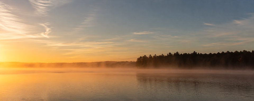 Sunrise over a still lake, mist drifting across the water in front of a dark treeline